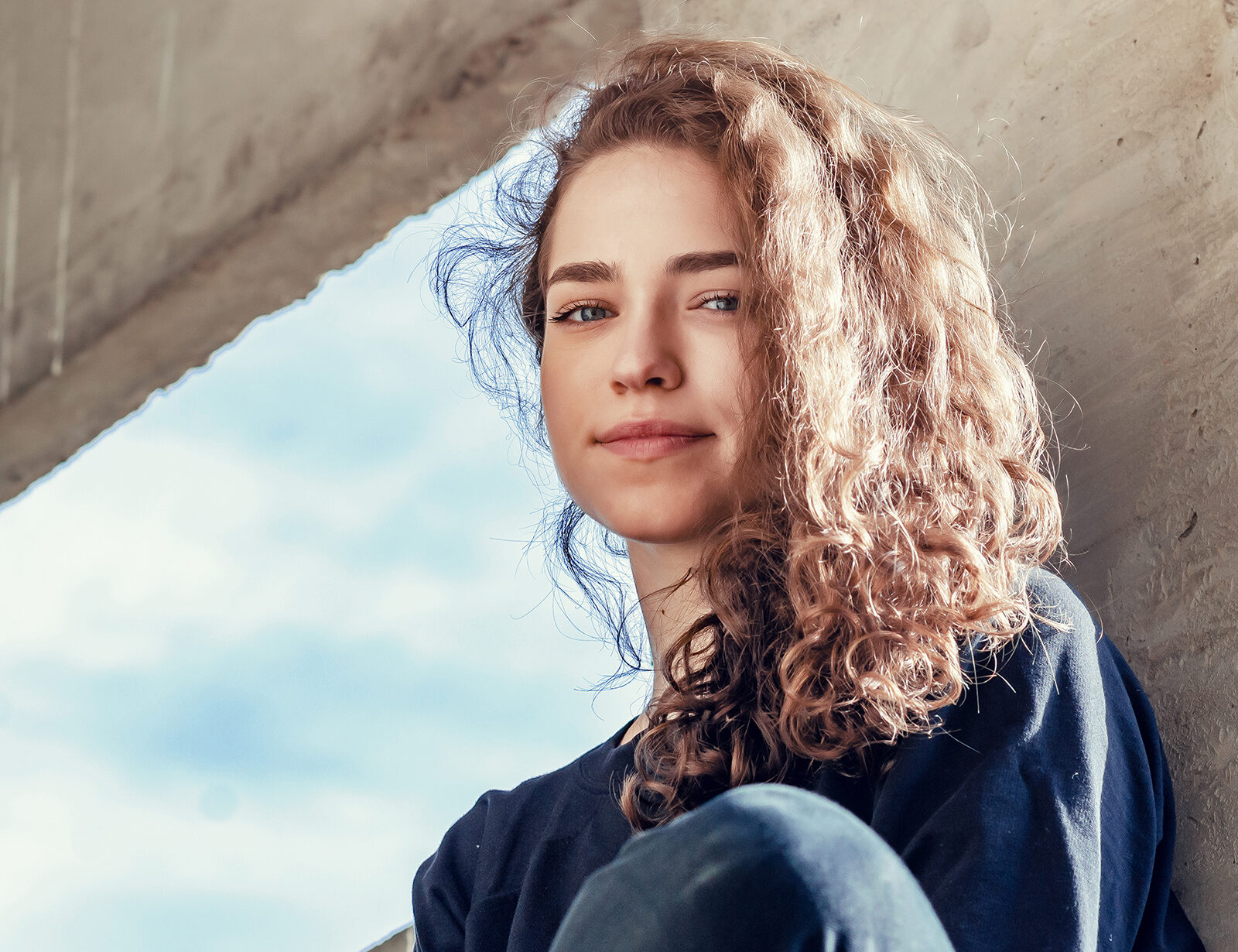 Lovely girl in dark clothes sits on concrete block in industrial
