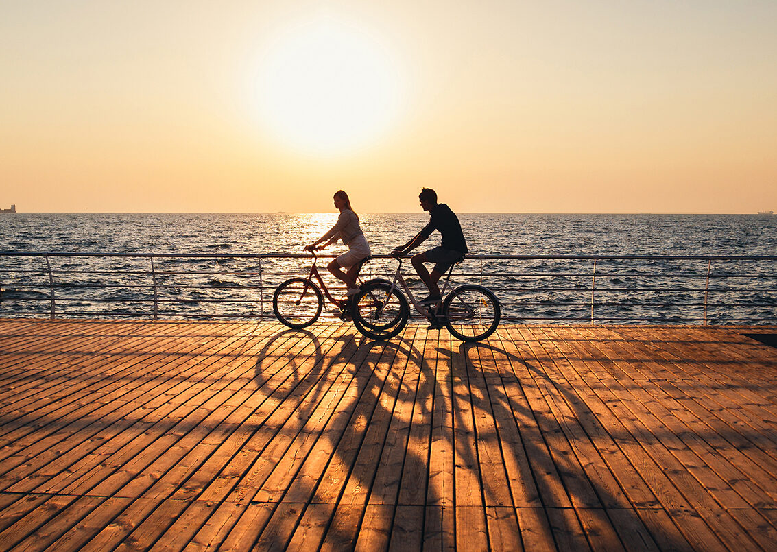 Couple of young hipsters cycling together at the beach at sunrise sky at wooden deck summer time