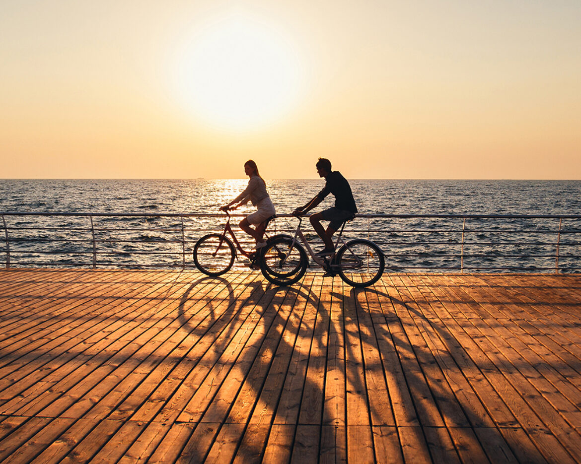 Couple of young hipsters cycling together at the beach at sunrise sky at wooden deck summer time