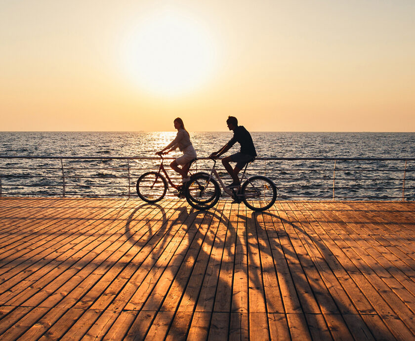 Couple of young hipsters cycling together at the beach at sunrise sky at wooden deck summer time